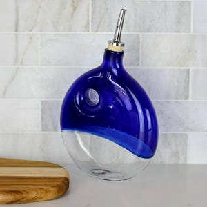 Puede incluir: Una aceitera de vidrio azul cobalto y transparente con un cuerpo redondeado y asimétrico que presenta un recorte circular cerca de la parte superior. El recipiente está rematado con un tapón de corcho y un vertedor de acero inoxidable.