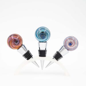 May include: Three glass bottle stoppers with colorful swirl designs. The stoppers are silver with black rubber bases and are displayed on a white stand.