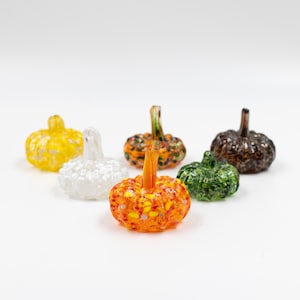 May include: A collection of six decorative glass pumpkins in various colors. The pumpkins are orange, yellow, white, green, brown, and orange with yellow speckles. Each pumpkin has a stem and a textured surface, perfect for fall decor.