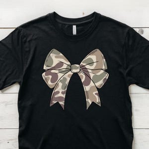 May include: A black short-sleeved t-shirt featuring a large, centered graphic of a bow. The bow is decorated with a camouflage pattern in shades of tan, brown, and olive green. The shirt has a crew neckline and a standard fit, with a visible brand label.