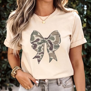 May include: A cream-colored short-sleeve t-shirt featuring a large, centered graphic of a bow with a camouflage pattern in shades of olive green, brown, and tan. The design includes bold black outlines, and the shirt has a classic crew neckline.