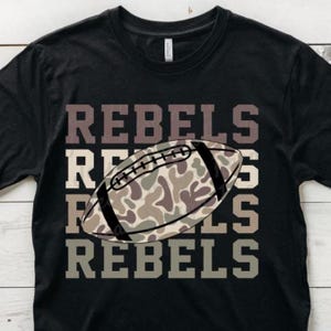 May include: Black t-shirt with the word "REBELS" stacked four times in a brown font. A football with a camouflage pattern is centered on the shirt. The shirt is made of a soft material.