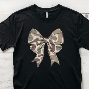 May include: A black short-sleeved t-shirt featuring a large, centered graphic of a bow. The bow is decorated with a camouflage pattern in shades of tan, brown, and olive green. The shirt has a crew neckline and a standard fit with visible stitching.