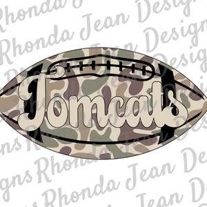 Peut inclure: Un ballon de football de couleur camouflage marron et beige avec le mot "Tomcats" en blanc.