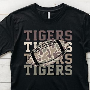 May include: Black t-shirt with the word "TIGERS" in a stacked design. A football with a camouflage pattern is centered on the shirt. The shirt is made of a soft material.