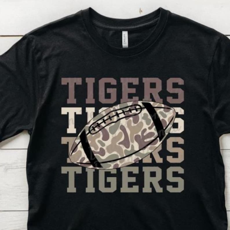 May include: Black t-shirt with the word "TIGERS" in a stacked design. A football with a camouflage pattern is centered on the shirt. The shirt is made of a soft material.