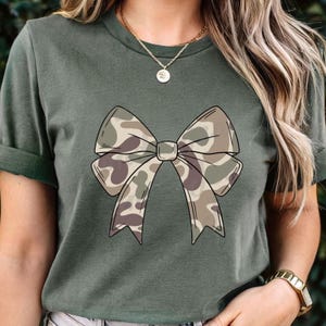May include: A dark olive green short-sleeve t-shirt featuring a large, centered graphic of a bow. The bow has a camouflage pattern in shades of tan, brown, and olive green. The shirt has a crew neckline and is worn with a gold-toned chain necklace.