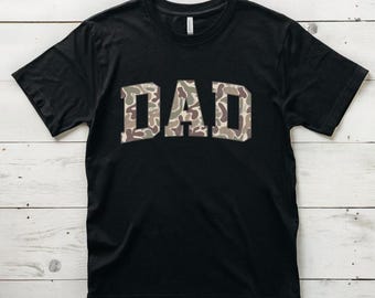 Op de afbeelding: Zwart T-shirt met het woord "DAD" in grote witte letters met een camouflagepatroon.