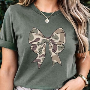 May include: A dark olive green short-sleeve t-shirt featuring a large, centered graphic of a bow. The bow has a camouflage pattern in shades of tan, brown, and olive green. The shirt has a crew neckline and is worn with a gold chain necklace.