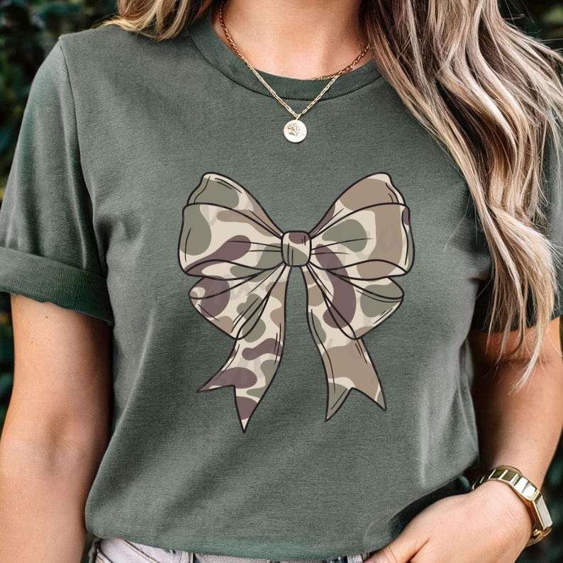 May include: A dark olive green short-sleeve t-shirt featuring a large, centered graphic of a bow. The bow has a camouflage pattern in shades of tan, brown, and olive green. The shirt has a crew neckline and is worn with a gold chain necklace.