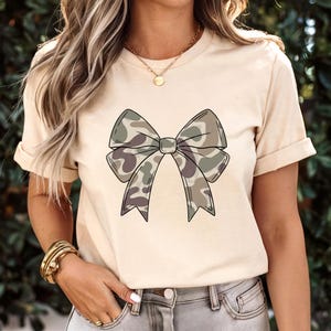 May include: A cream-colored short-sleeve t-shirt featuring a large, centered graphic of a bow with a camouflage pattern in shades of olive green, brown, and tan. The bow has a distinct black outline and is positioned on the front of the shirt.