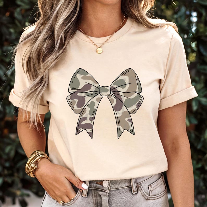 May include: A cream-colored short-sleeve t-shirt featuring a large, centered graphic of a bow with a camouflage pattern in shades of olive green, brown, and tan. The bow has a distinct black outline and is positioned on the front of the shirt.