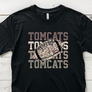 May include: Black t-shirt with the word "TOMCATS" stacked four times in a gradient of brown and gray. A football with a camouflage pattern is centered on the shirt.