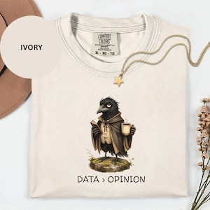 Pode incluir: Uma t-shirt cor de marfim com um gráfico de um corvo preto a usar uma capa, segurando um livro e uma chávena de café enquanto está de pé sobre uma pedra. Abaixo do gráfico, o texto diz DATA > OPINION. A t-shirt tem uma etiqueta Comfort Colors.