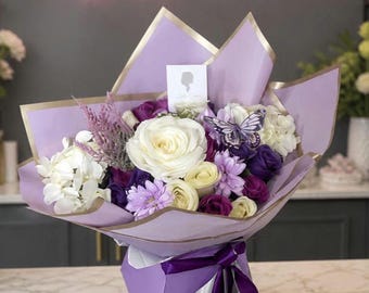 May include: A bouquet of white and purple flowers including roses and hydrangeas, wrapped in lavender paper with gold trim and a purple ribbon. A decorative butterfly and a small white card with the text Lady Grace Flowers are included.