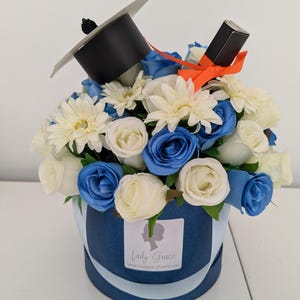 May include: A blue and white flower arrangement in a blue box with a graduation cap and a black and orange ribbon on top. The box has a label that says "Lady Grace" and "www.ladygraceflowers.com".