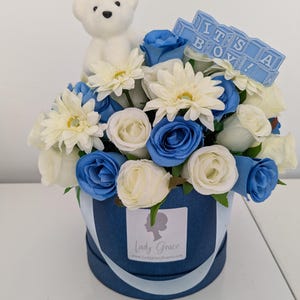 May include: A blue and white flower arrangement in a blue hatbox with a white teddy bear and blue blocks that spell "It's a Boy!"