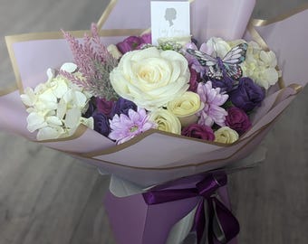 Könnte beinhalten: Ein Strauß weißer und lila Blumen, die in einer lila Box mit einem lila Band angeordnet sind. Die Blumen sind in helllila Papier mit goldener Umrandung verpackt. Eine kleine Karte mit dem Text "Lady Grace" ist am Strauß befestigt.