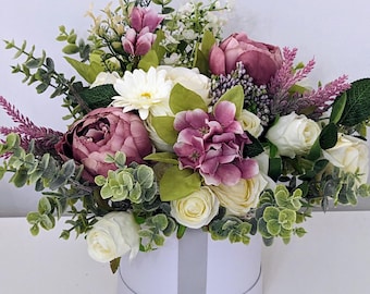 Könnte beinhalten: Eine weiße Box gefüllt mit einem Strauß künstlicher Blumen. Die Blumen sind eine Mischung aus weißen, rosa und violetten Rosen, Hortensien und anderen Blüten. Die Box hat ein graues Band darum.
