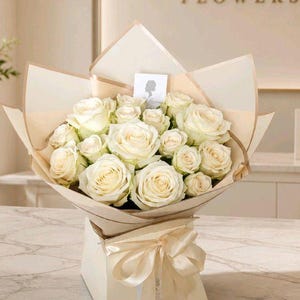 May include: A bouquet of cream-coloured roses arranged in a square box with a matching ribbon. The flowers are wrapped in beige paper with a small white card featuring a silhouette logo. The arrangement is displayed in a floral shop setting.