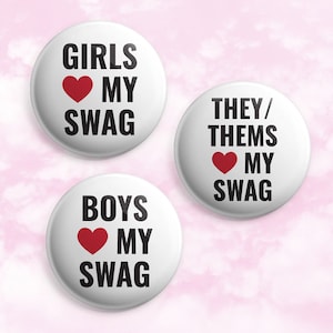 May include: Three white button pins with black text and a red heart. The pins read "Girls love my swag", "Boys love my swag", and "They/Them love my swag".