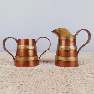 May include: Two vintage copper and brass pitchers. The pitchers have a copper body with brass bands and handles. The pitcher on the right has a spout. The pitchers are on a cream-colored, textured surface.