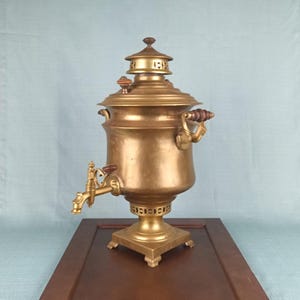 May include: A vintage brass samovar with a spigot, handles, and a decorative base. The tea kettle has a domed lid and a warm, golden hue. The samovar is displayed on a dark brown wooden surface.