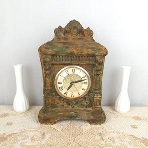 May include: A vintage wooden clock with a distressed finish, featuring a round clock face with Roman numerals. The clock is flanked by two white vases. The clock has a decorative, ornate design and is likely a mantel clock.