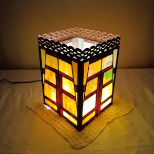 May include: A stained glass lamp with a square shape, featuring yellow, red, and green glass panels. The lamp has a black metal frame and a decorative scalloped edge. It sits on a cream-colored, crocheted doily, and is illuminated from within.