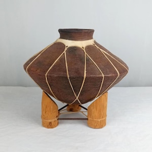May include: A handcrafted, brown, geometric-shaped vessel with a light-colored rim and decorative string accents. The vessel is supported by a wooden stand with three legs. The vessel is likely used for decorative purposes.