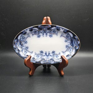 Puede incluir: Un plato ovalado de cerámica con un centro blanco y un borde decorativo con intrincados patrones florales en azul oscuro y azul claro. El borde está acentuado con finas líneas doradas y la pieza se exhibe sobre un soporte de madera.