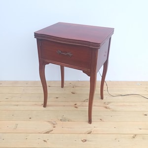 May include: A dark brown wooden sewing machine cabinet with a rectangular top, a single front drawer featuring a decorative metal handle, and four curved legs. A thin black power cord extends from the side of the wooden furniture piece.