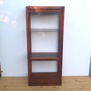May include: A tall, rectangular wooden shelving unit with a medium-brown wood grain finish. The structure features a solid top and base, with one clear glass shelf positioned in the upper section and a solid wooden shelf in the middle section.
