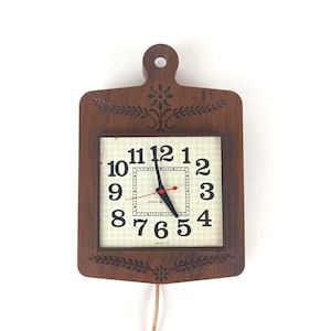 Puede incluir: Un reloj de pared de madera vintage con forma de tabla de cortar. La esfera del reloj es cuadrada con grandes números negros y un segundero rojo. El reloj es marrón con tallas decorativas de flores y hojas.