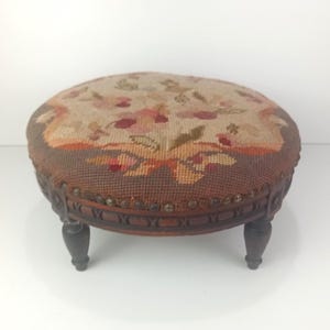Puede incluir: Un reposapiés redondo con una parte superior de punto de aguja con un patrón floral en tonos rosa, verde y bronceado, bordeado por una tela marrón oscuro. El marco de madera tiene detalles tallados y se apoya en cuatro patas torneadas.