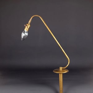 May include: A vintage brass floor lamp with a curved arm and a clear, teardrop-shaped lightbulb. The lamp has a round base and a slender, vertical pole. The overall design is minimalist and elegant, suitable for a living room or bedroom.