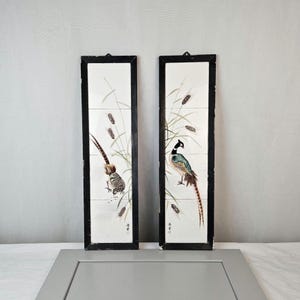May include: Two framed wall art panels featuring detailed illustrations of pheasants amidst reeds. The birds display a variety of colors, including brown, green, and blue, set against a white background. The frames are black, adding a contrasting border to the artwork.