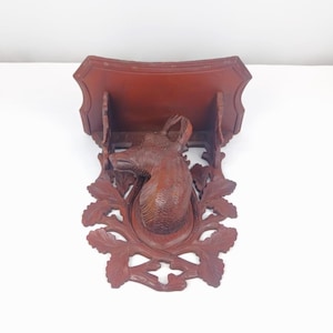 May include: A carved wooden wall shelf with a deer head design. The shelf is a dark reddish-brown color and features a detailed carving of a deer head in the center, surrounded by decorative leaves. The shelf has a rectangular shape.