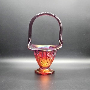 Op de afbeelding: Een mand van carnavalsglas met een dieprode en oranje iriserende afwerking. Het lichaam vertoont een geperst ruitpatroon met een geschulpte rand en een hoog, gebogen handvat. De basis is zeshoekig en ondersteunt de uitlopende, textuurrijke kom.