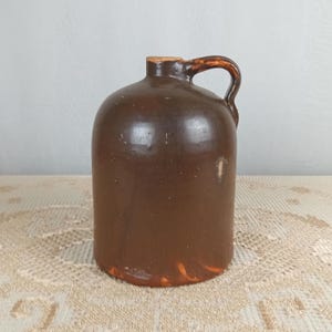 May include: A brown stoneware jug with a handle. The jug has a rounded body and a narrow neck. The handle is arched and attached to the top of the jug. The jug is made of a dark brown clay and has a glossy finish. The jug is likely an antique.