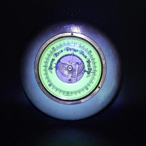 May include: A round, metallic barometer with a glowing light green face. The dial features the text Stormy, Rain, Change, Fair, and Very Dry, along with numerical markings from 1 to 31. A metal ring is attached to the top of the circular frame.