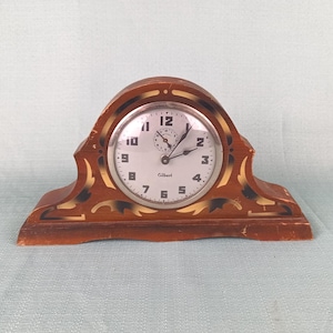 May include: A vintage Gilbert clock with a wooden frame. The clock face is white with black numerals and hands. The frame is brown with gold and black decorative accents. The clock is a classic, decorative piece.