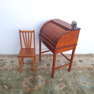 May include: A miniature wooden roll-top desk with a matching small wooden chair. The desk features a curved, slatted top and a single drawer, while the chair has a fan-shaped backrest. Both pieces are crafted from a medium-toned wood.