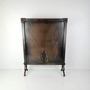 May include: A rectangular fireplace screen made of dark bronze-toned metal. The screen features a central fan-shaped motif with radiating lines extending towards the top corners. It is supported by four curved feet and has decorative corner accents.