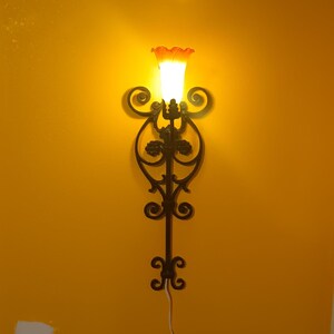 May include: A black wrought iron wall sconce with an orange and white glass shade. The light is on and illuminating the wall.