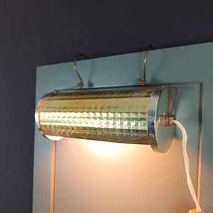 May include: A vintage-style wall-mounted light fixture. The cylindrical lamp has a textured, gold-colored metal shade with a silver trim. It is attached to a light blue surface with a white cord and a hook for hanging.