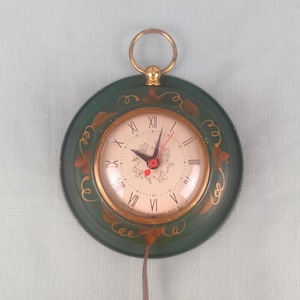 May include: A vintage wall clock with a dark green circular frame and a gold-colored ring for hanging. The clock face is cream-colored with black Roman numerals and red hands. Gold-colored leaf designs adorn the frame. The clock reads "CLOCK MOVEMENT BALTIMORE MADE IN U.S.A."