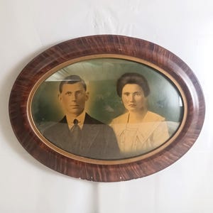 Puede incluir: Una fotografía antigua de forma ovalada en un marco de madera oscura. La imagen en tonos sepia muestra a un hombre y una mujer, probablemente una pareja, con un fondo verde. El marco tiene un rico patrón de vetas arremolinadas.