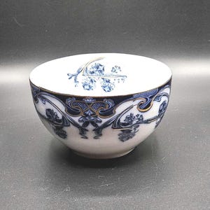 May include: A white ceramic bowl featuring a dark blue and gold decorative pattern around the exterior. The interior of the bowl displays a blue floral design at the bottom. The surface of the bowl shows a fine crackle finish throughout the glaze.