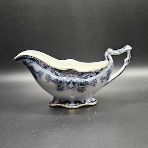 Puede incluir: Una salsera de cerámica con base blanca y detallados patrones florales y de volutas en azul oscuro. El recipiente cuenta con un asa curva, un pico vertedor ancho y una base festoneada, todo acentuado con un delicado borde dorado.